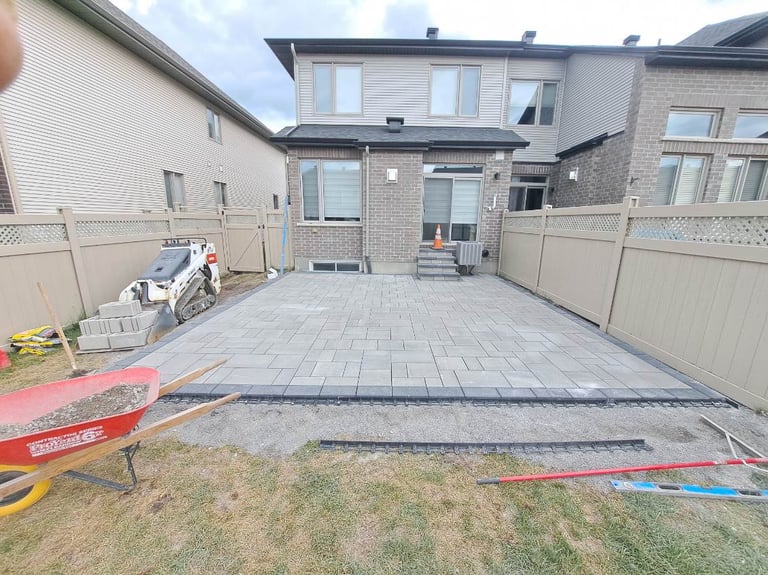 New paver patio in residential backyard with brick home, construction equipment, and colored marking lines on grass