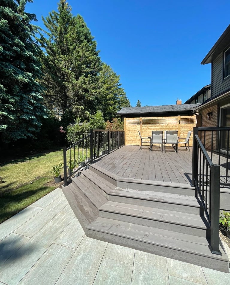 Multi-level wooden deck with black railings extending from a modern house, surrounded by tall green trees under clear blue sky
