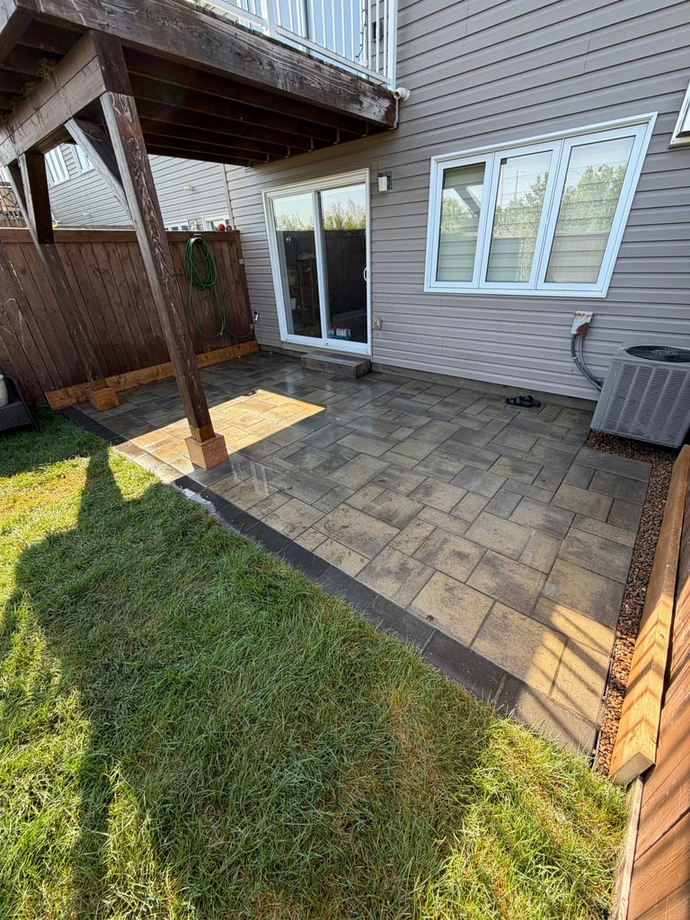 Backyard patio with gray stone pavers extending from sliding glass doors, wooden pergola on left, gray vinyl siding house, and green lawn