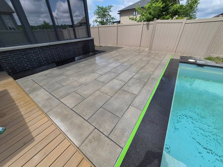 Modern residential patio with concrete pavers, wooden deck, pool edge with green border, and black brick house wall with windows