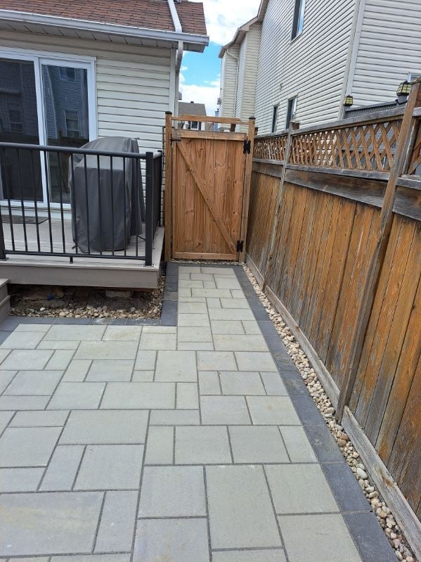 Residential backyard patio with gray stone pavers, wooden fence gate, metal deck railing, and two houses visible